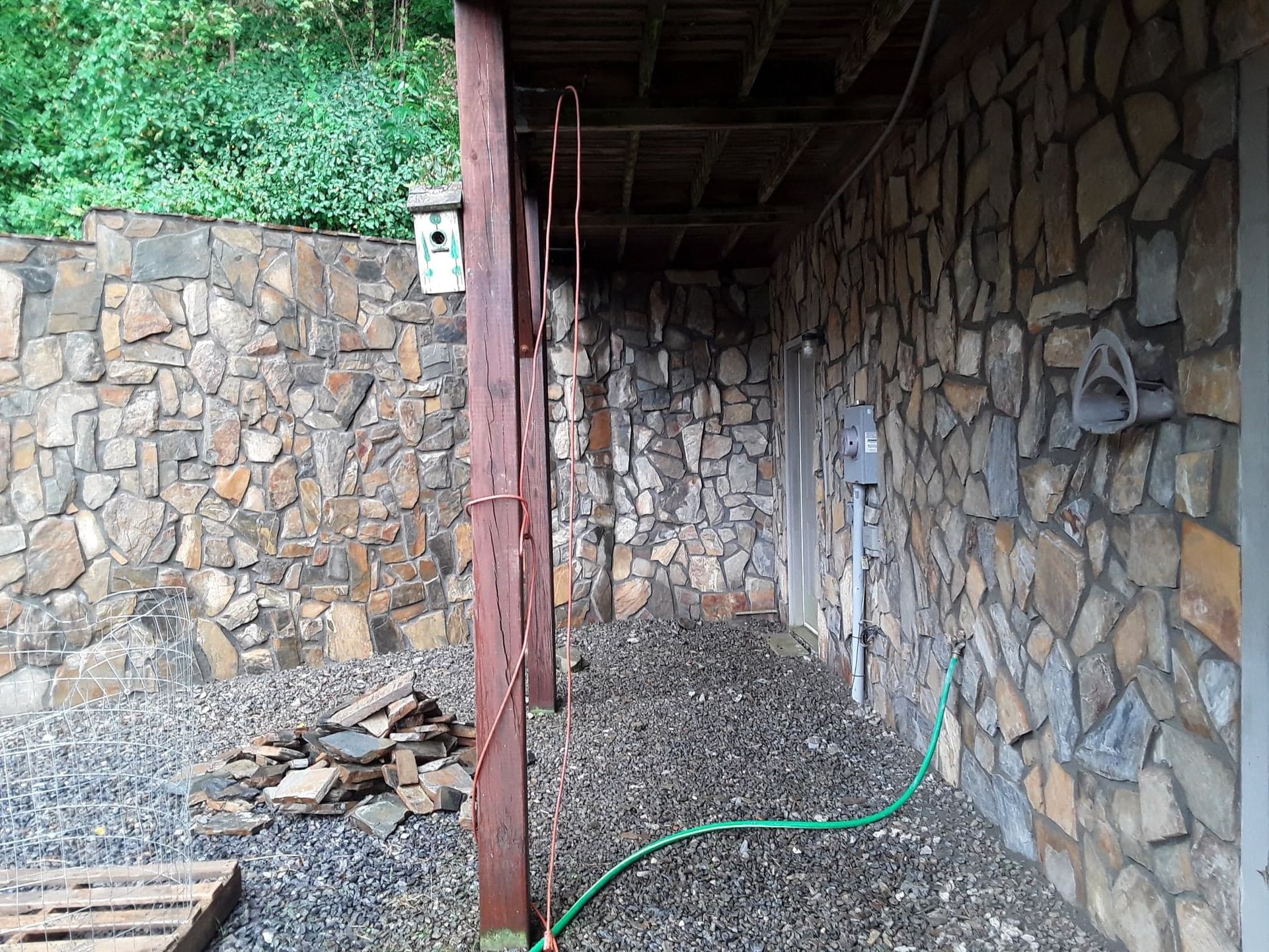 Stone walls with a covered walkway. A green hose, electrical box, and birdhouse are visible.