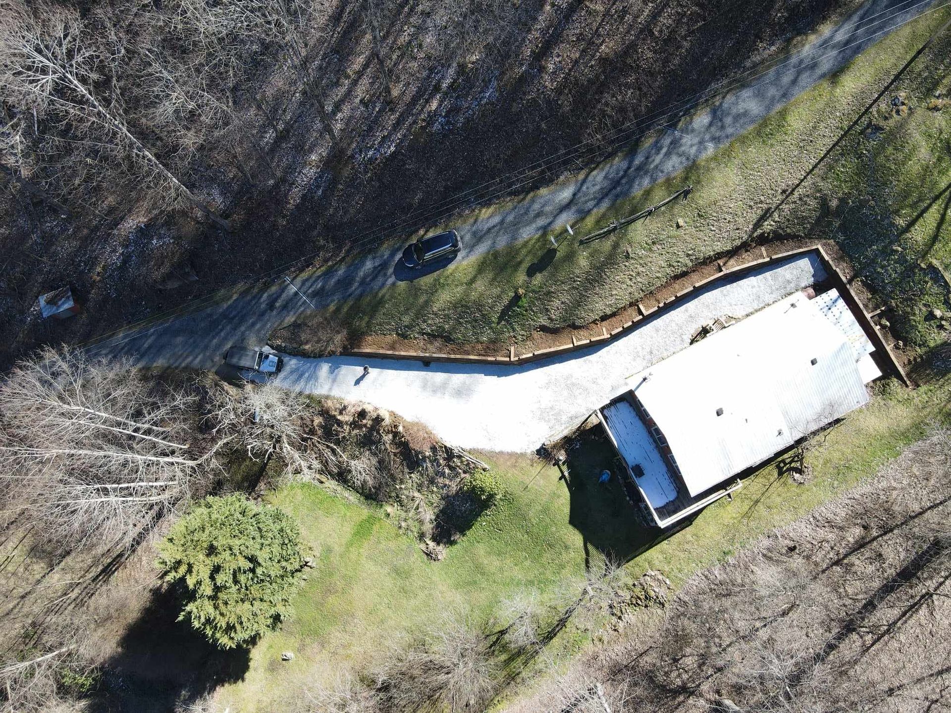 Overhead view of a house with a white roof next to a paved driveway and surrounded by grassy and wooded areas.