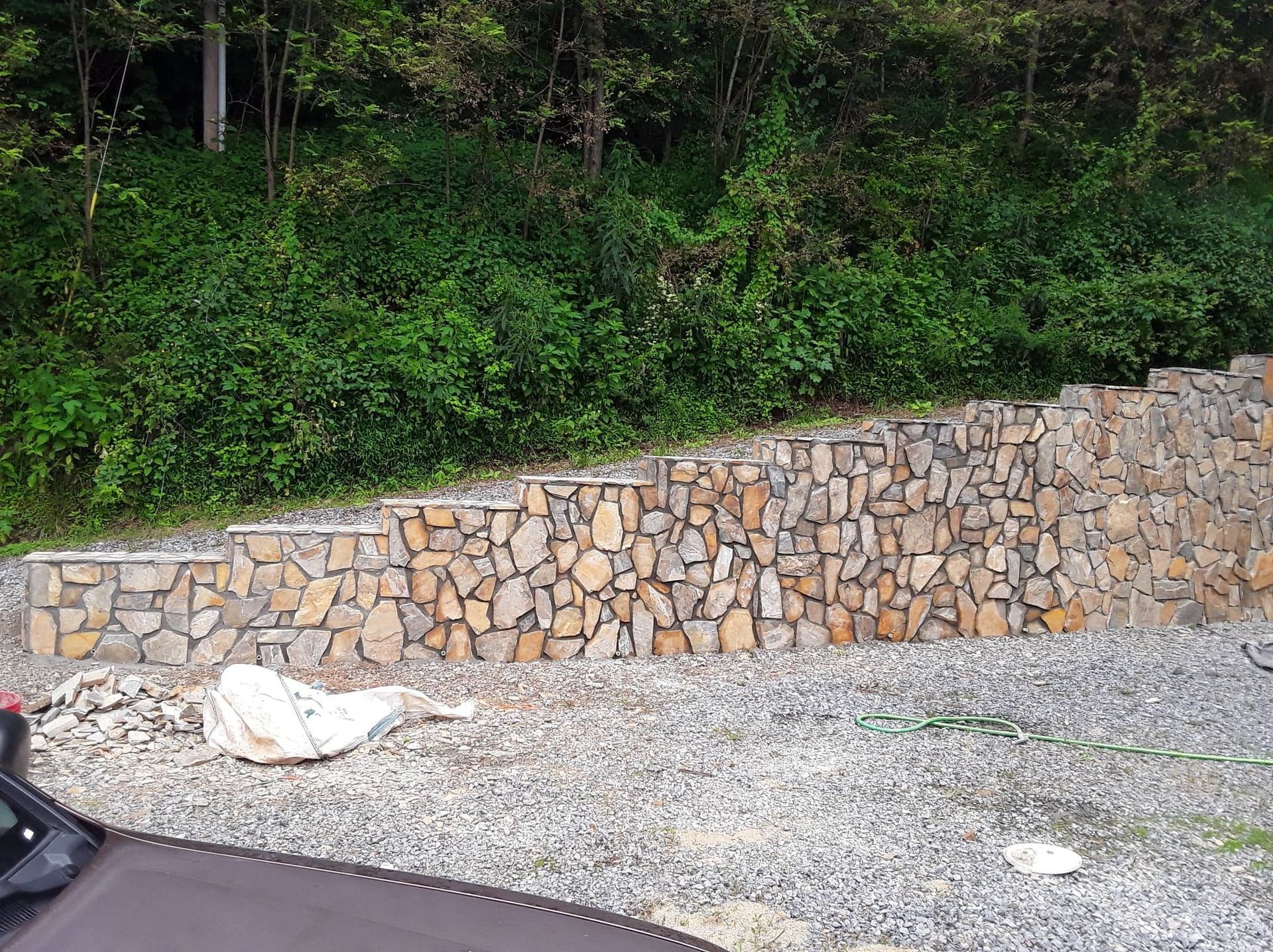 Stone retaining wall with steps, built into a hillside with lush green foliage. Gray gravel ground and a car in the foreground.