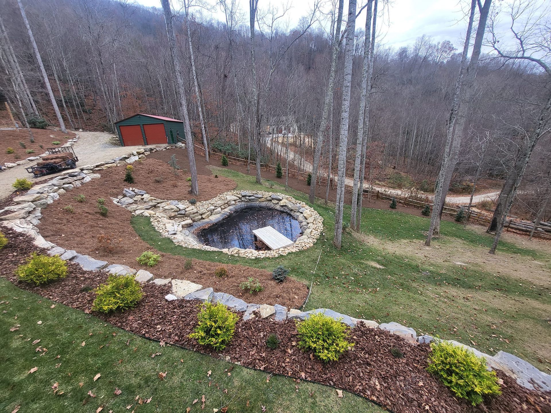 A backyard pond with a small wooden dock, surrounded by landscaping, trees, and a distant red shed.