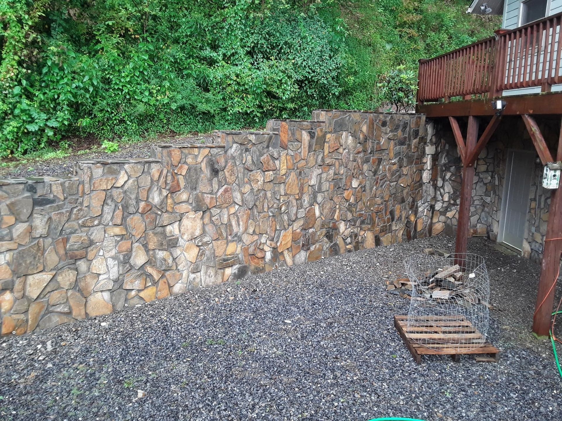 Stone retaining wall beside a wooden deck, with dark gravel ground and greenery above.