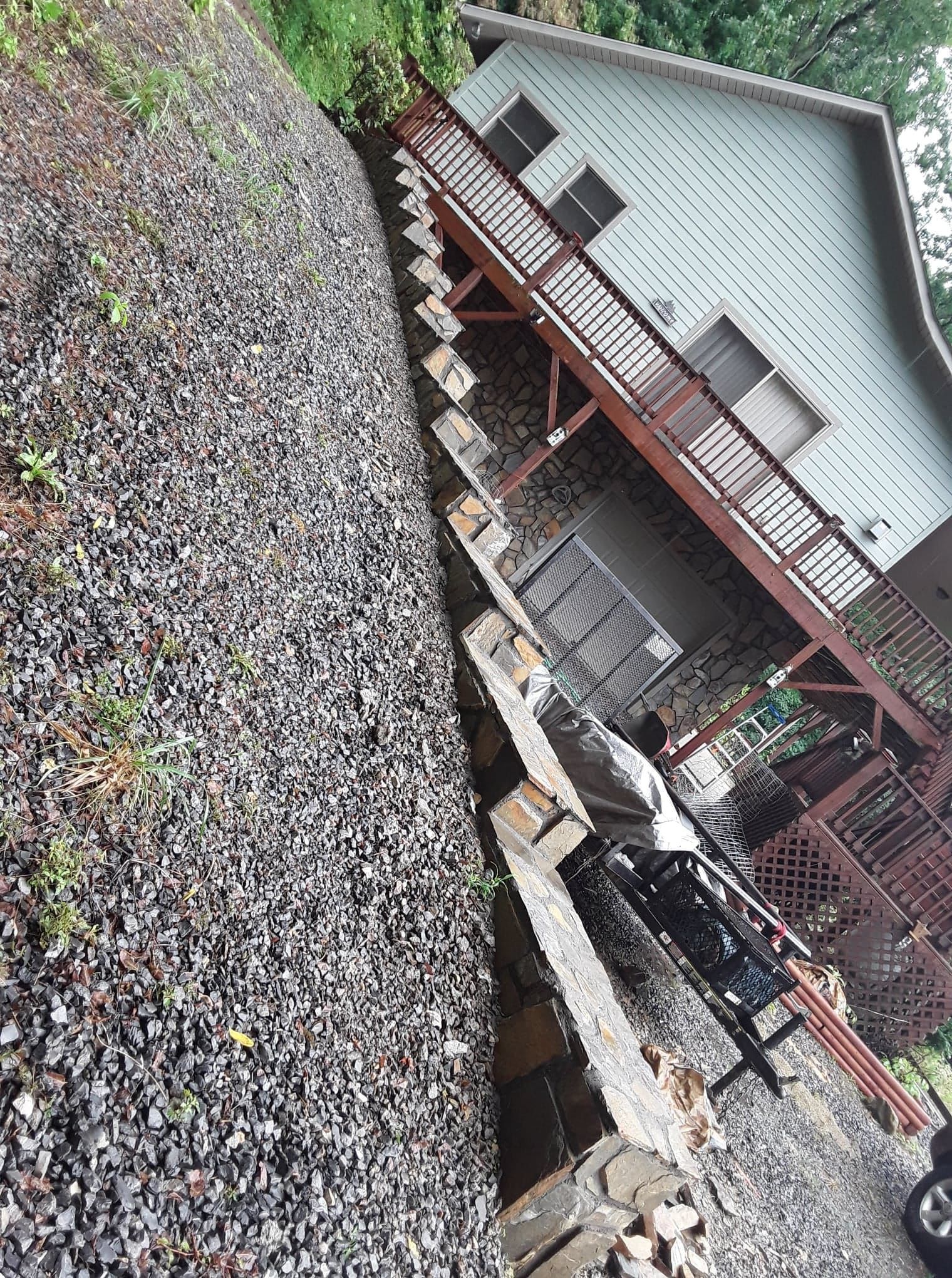 A person is standing on stone steps leading to a house. The steps are surrounded by gravel on a sloped hillside.