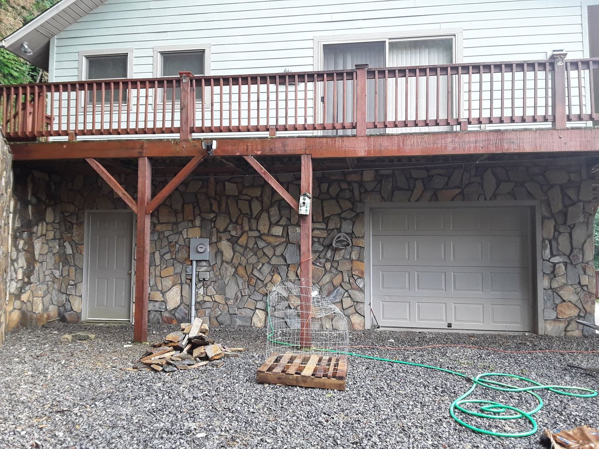 House with stone facade, wooden deck, and garage. A water hose and a pallet are in the foreground.