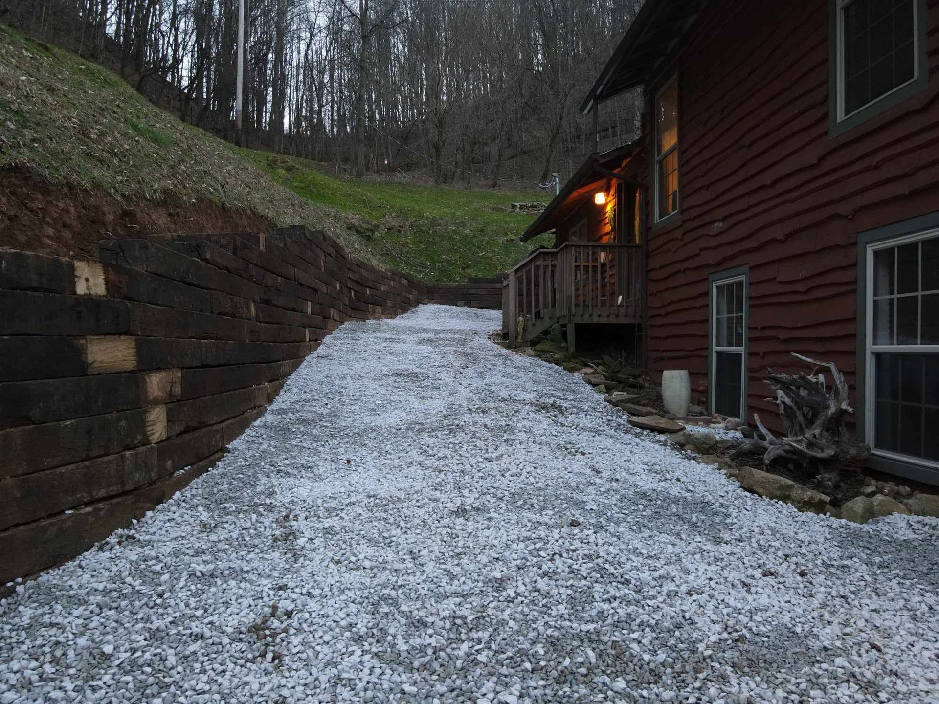 A gravel driveway leads up a hill past a wooden retaining wall and a reddish-brown house with lit windows.