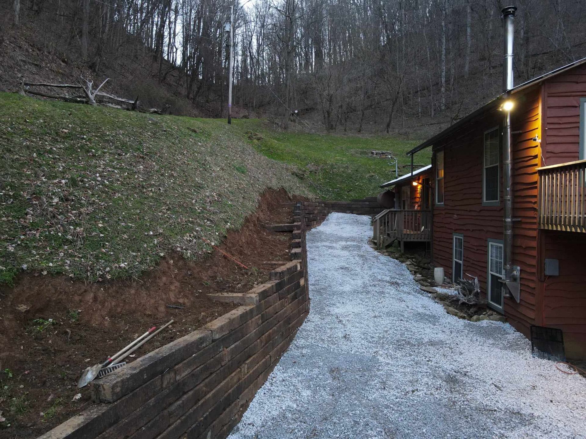 A gravel driveway leads to a wooden cabin in a hillside setting. A retaining wall is on the left.