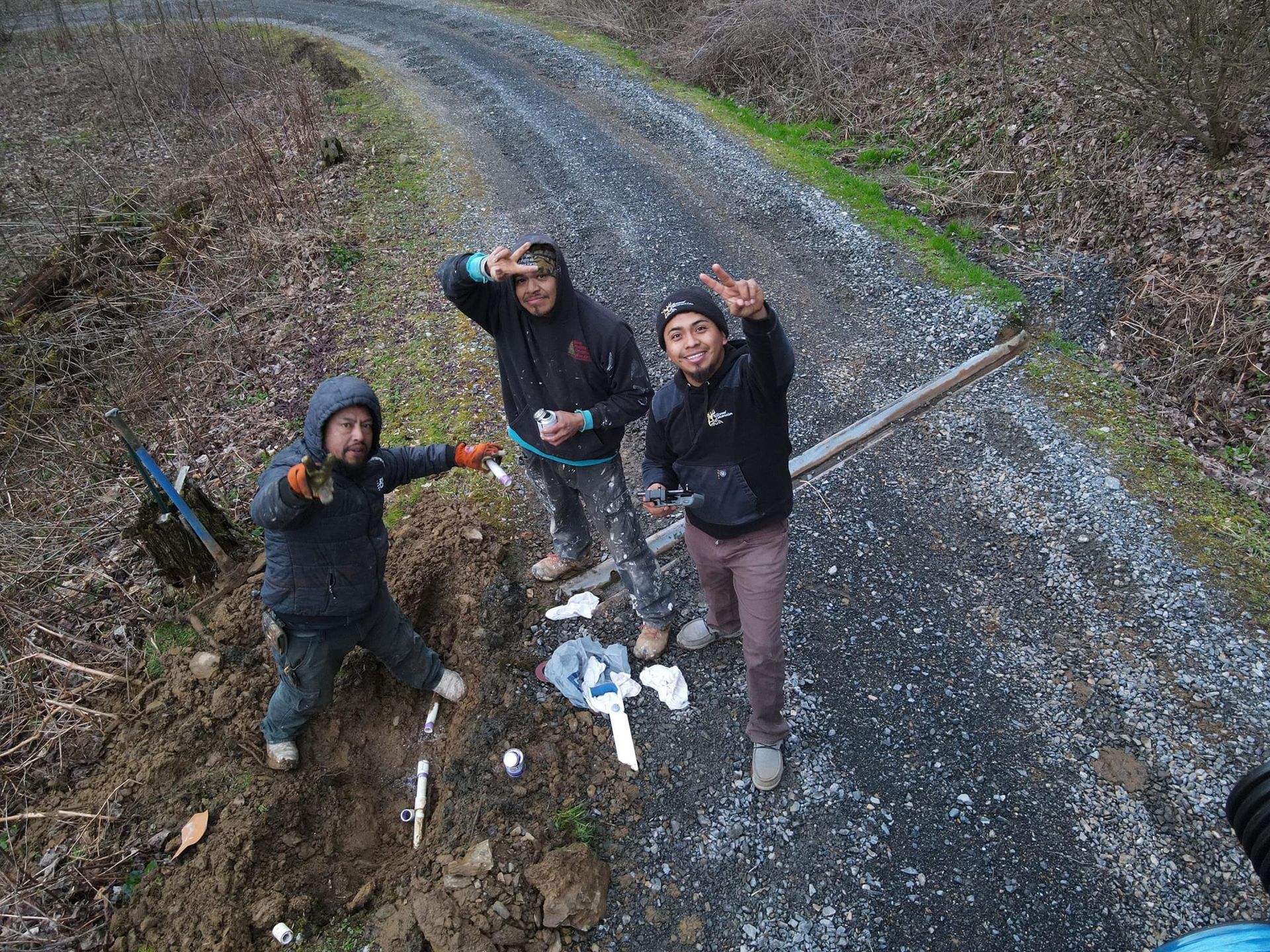 Three men outdoors on a gravel road, waving and smiling. One points, while another shades his eyes. Dark, cloudy day.
