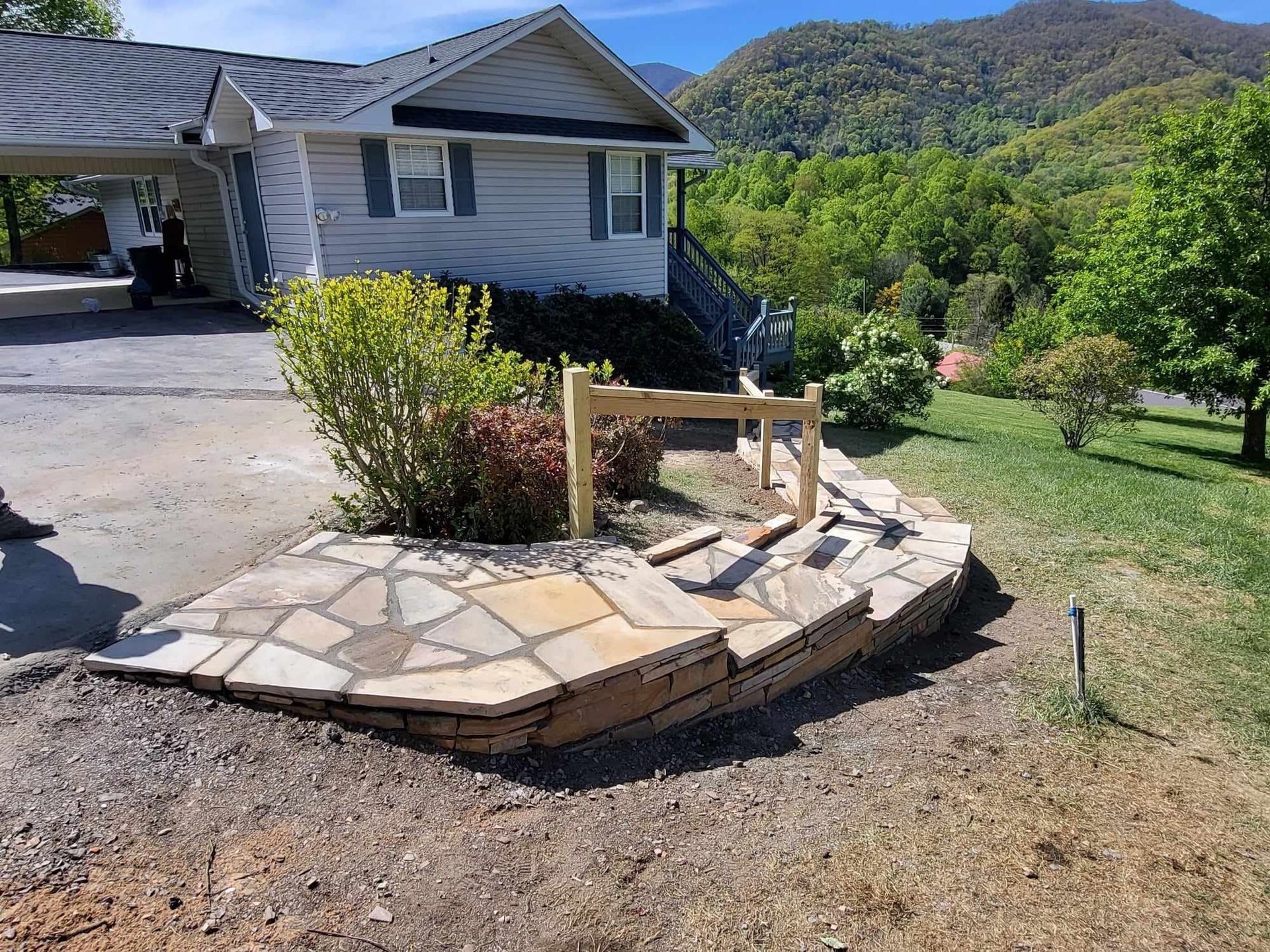 Stone-walled garden bed in front of a house; unfinished wooden frame and various plants. Mountains and trees in background.
