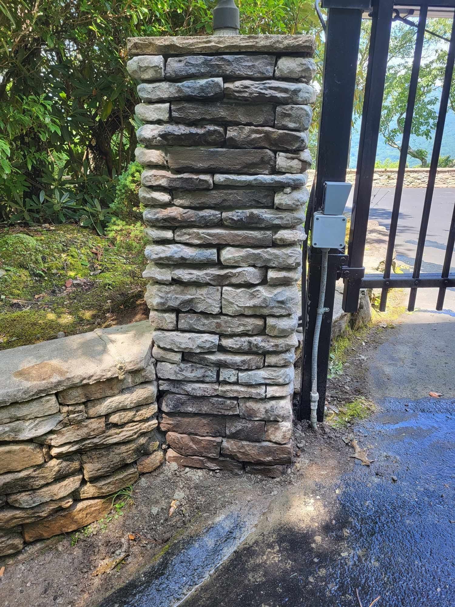 Stone pillar with a security gate, attached to a low stone wall. The pillar is grey and tan, and the gate is black.