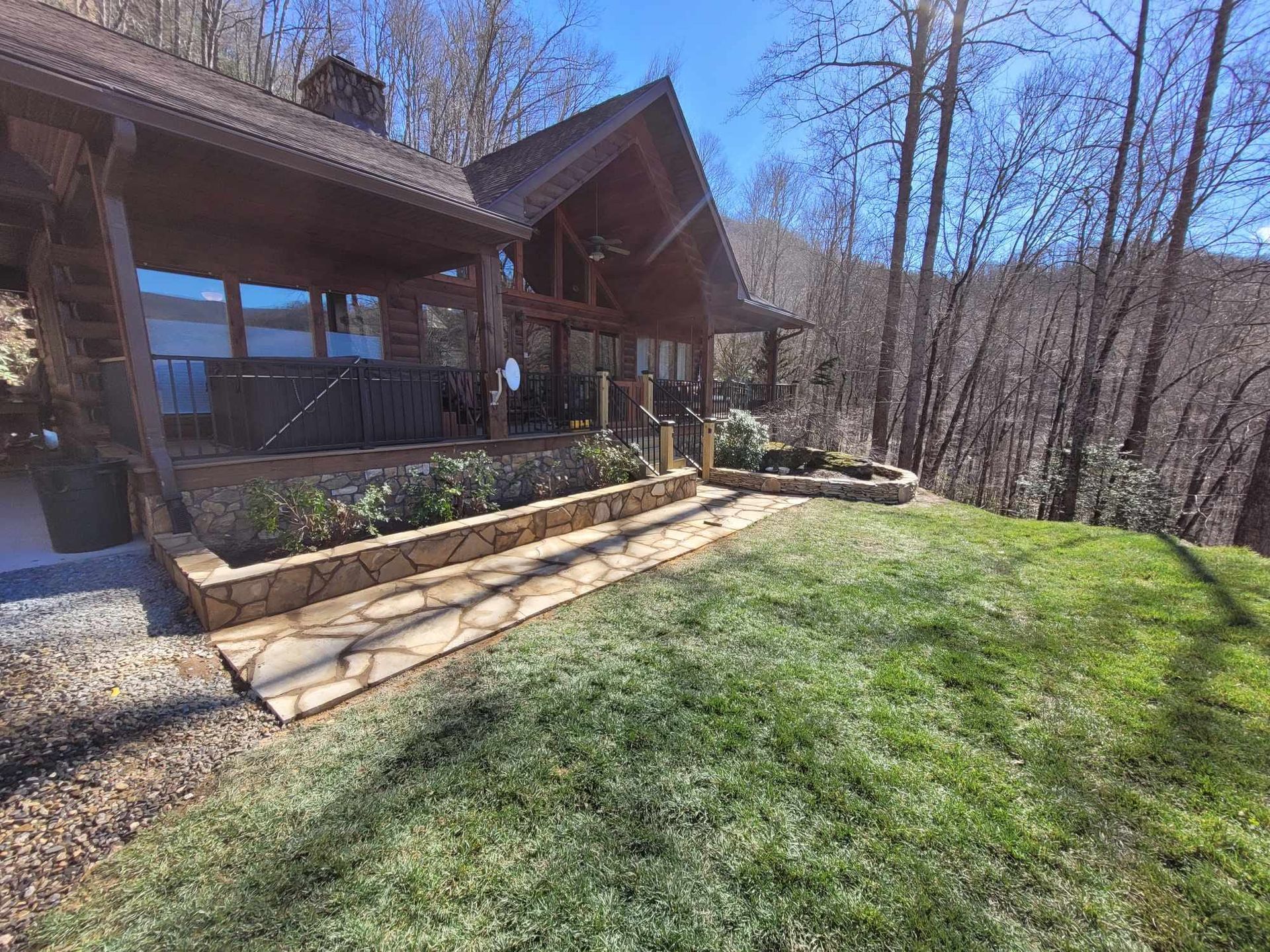 Wooden cabin with stone retaining walls and a lawn, set against a backdrop of trees under a blue sky.