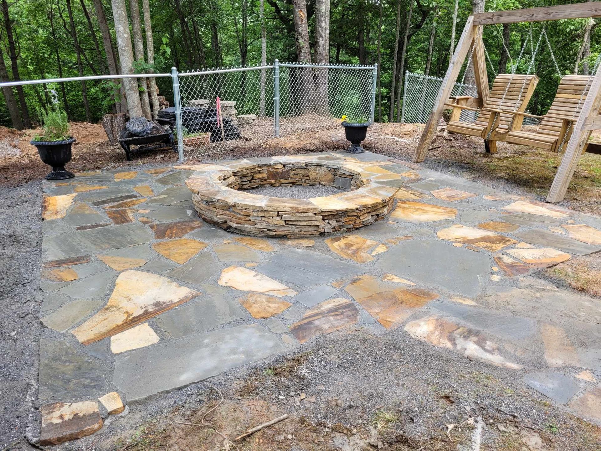 A stone fire pit in a backyard patio surrounded by flagstone, with wooden swing chairs visible.
