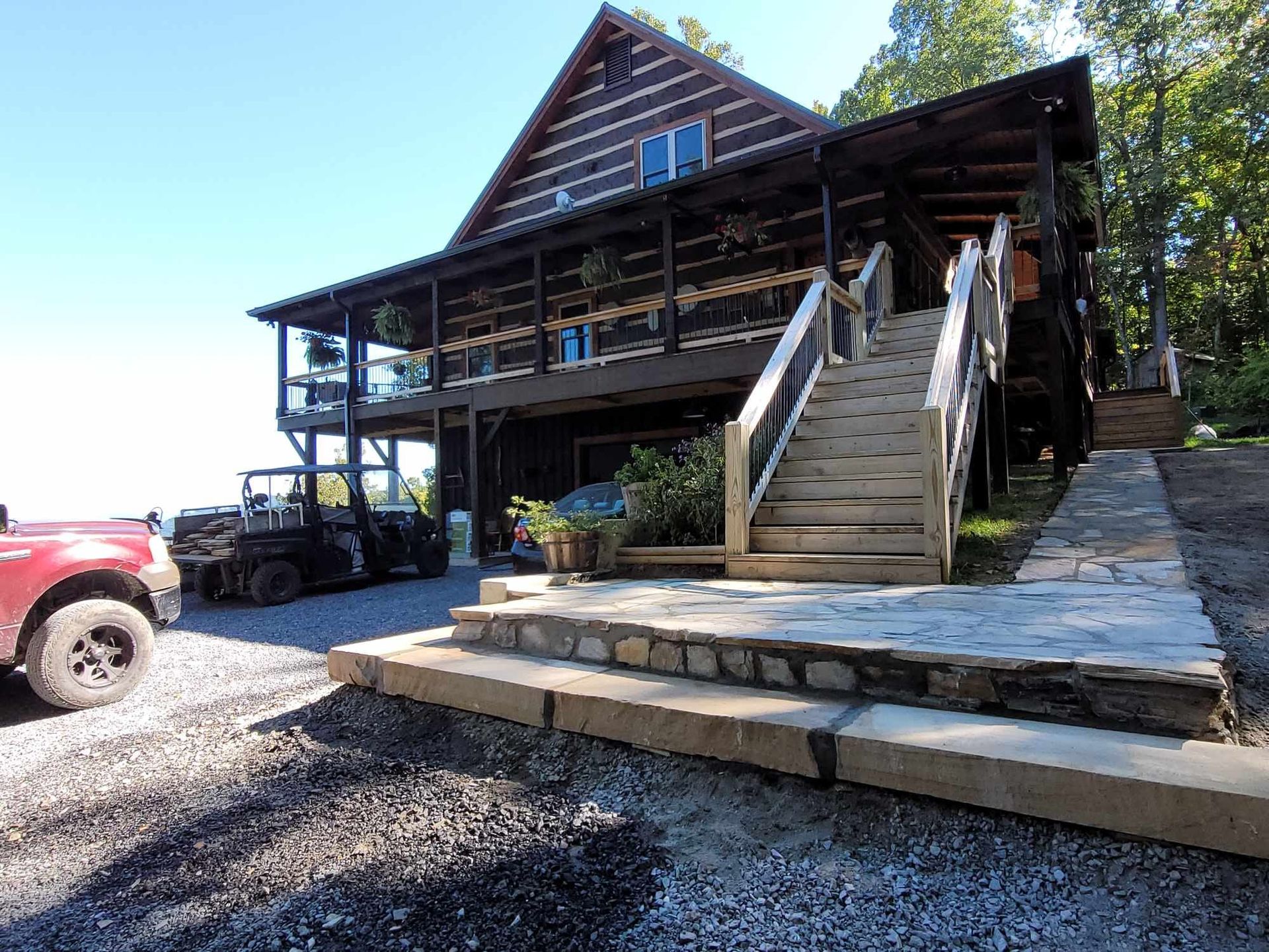 Log cabin with a large deck, steps, and a gravel driveway. A red truck and utility vehicle are parked nearby.