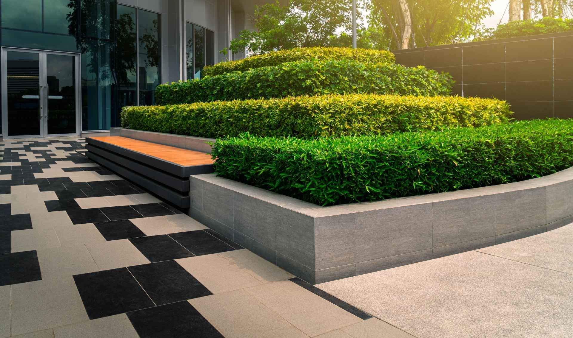 Modern building exterior with tiered, manicured hedges and built-in wooden bench, set on a patterned paved area.