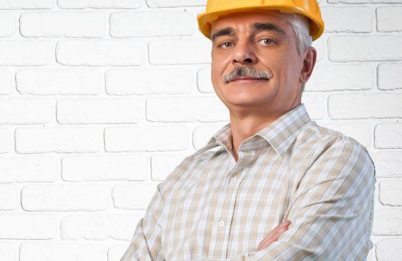A man wearing a hard hat is standing with his arms crossed in front of a white brick wall.