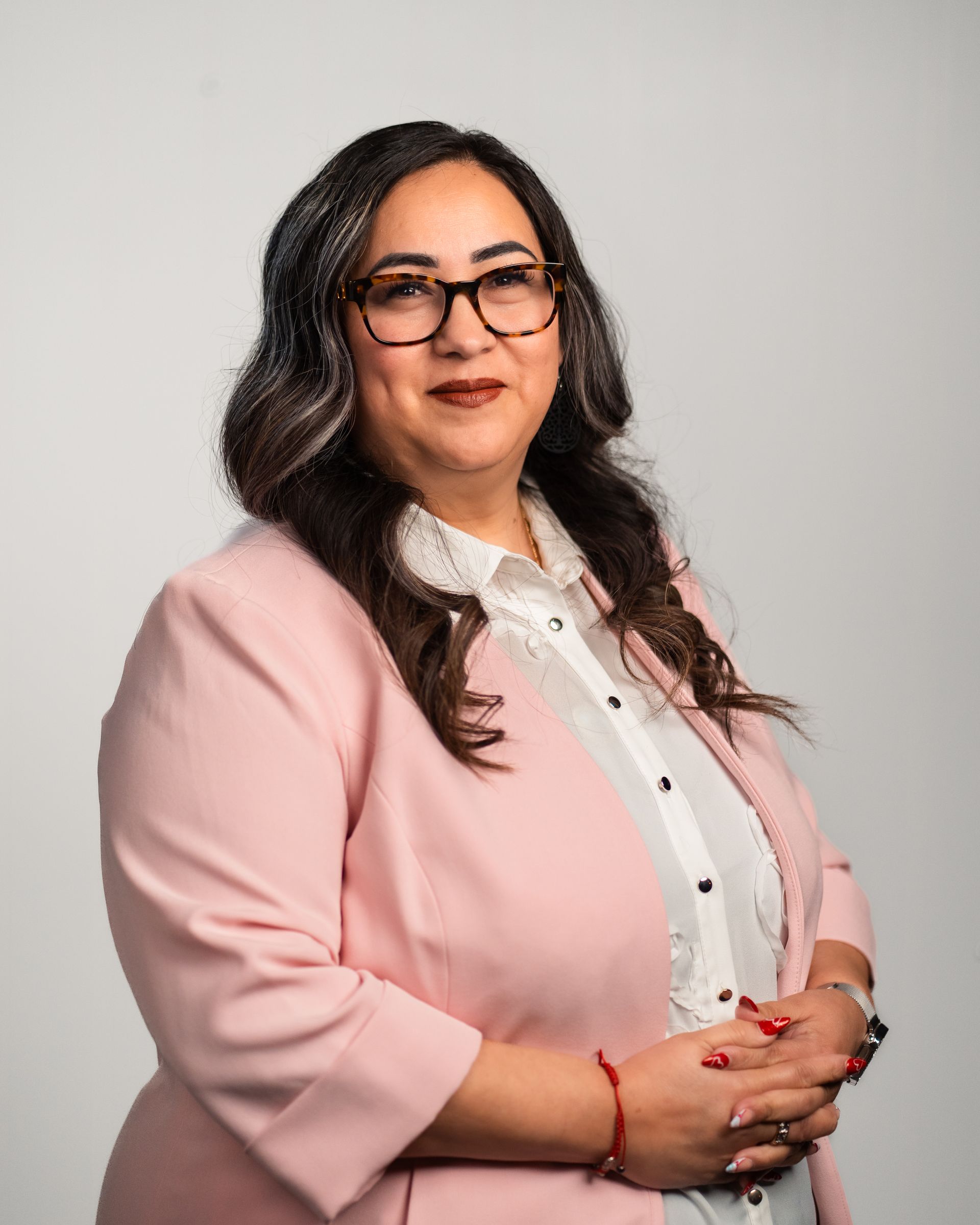 Adriana Villalobos wearing glasses and pink blazer, smiles, arms crossed, white background.