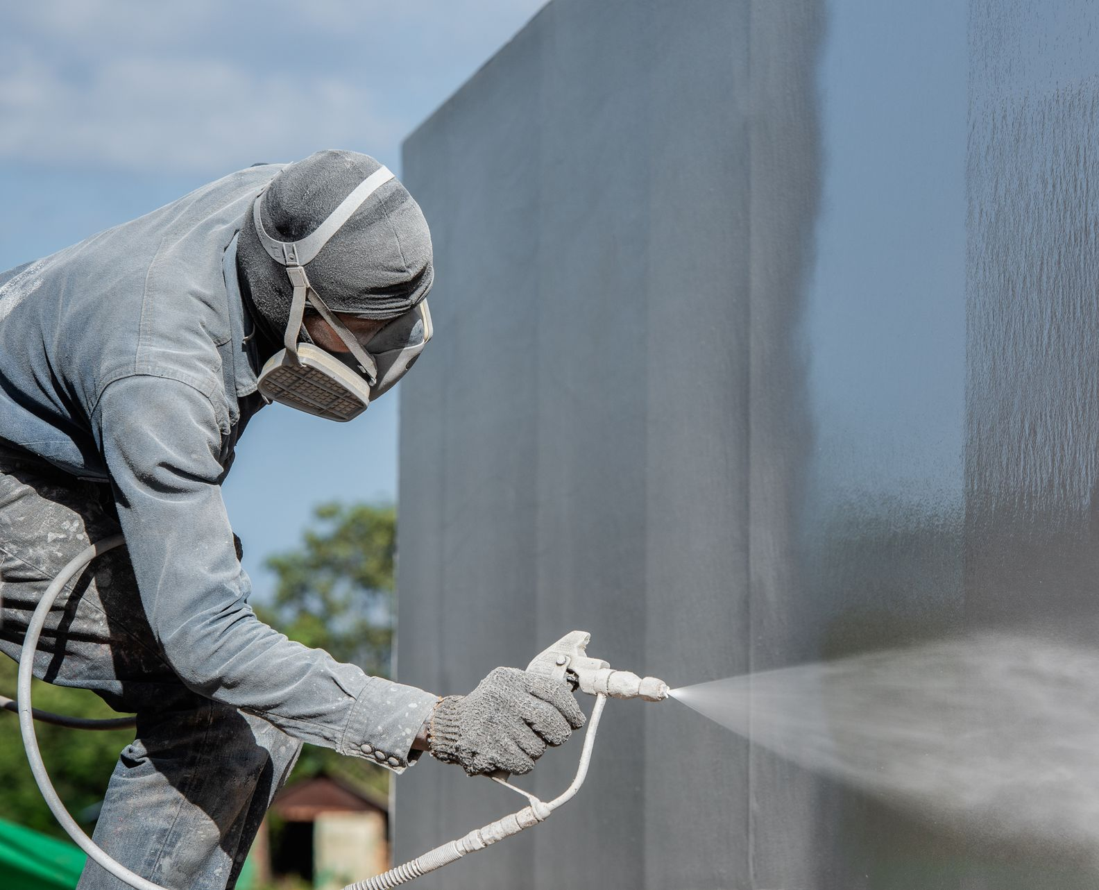 A person wearing a white cap and blue overalls uses a roller to paint a white section on a beige exterior wall.