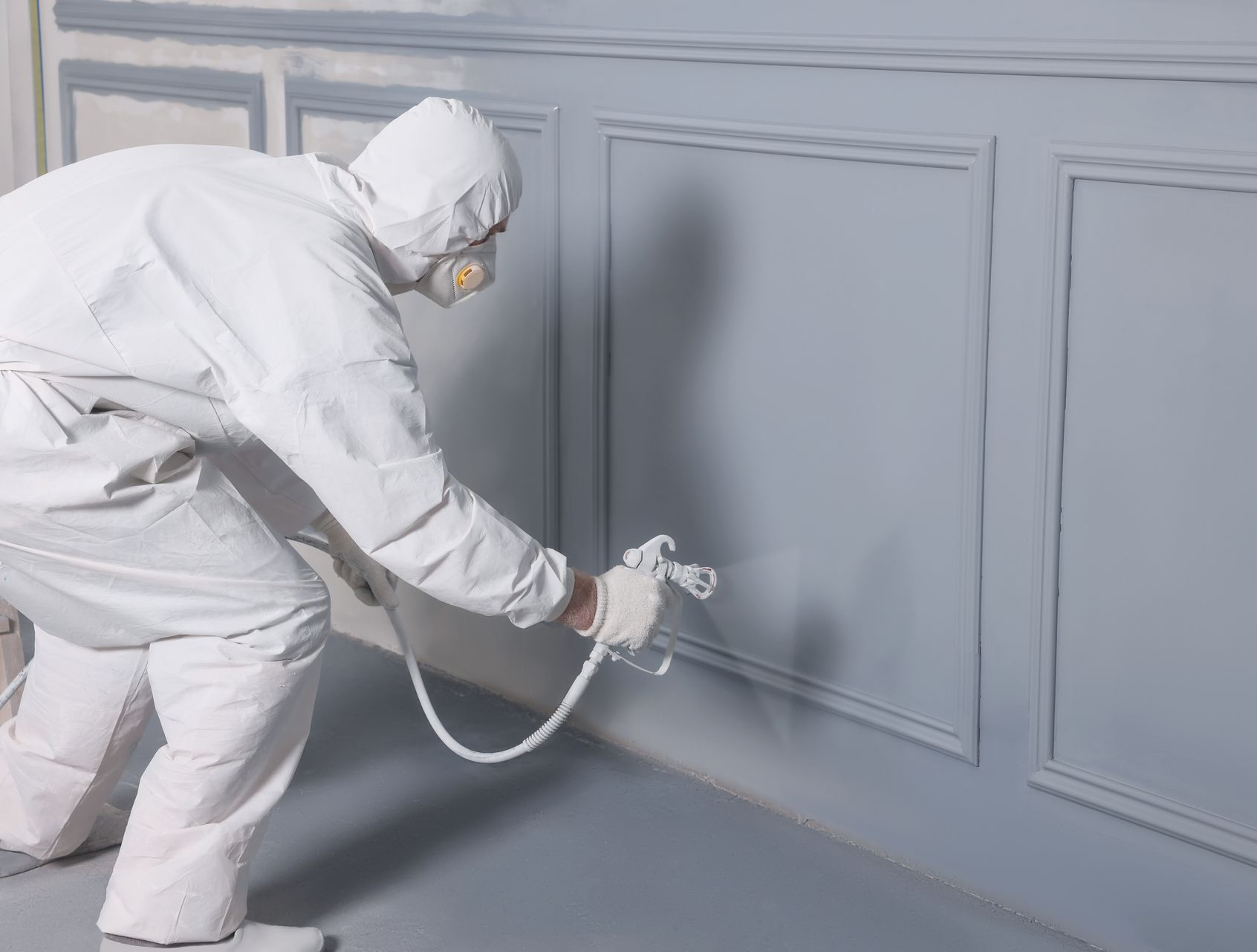 A person in a blue jumpsuit and hat uses a paint roller to apply white paint to a beige house exterior.