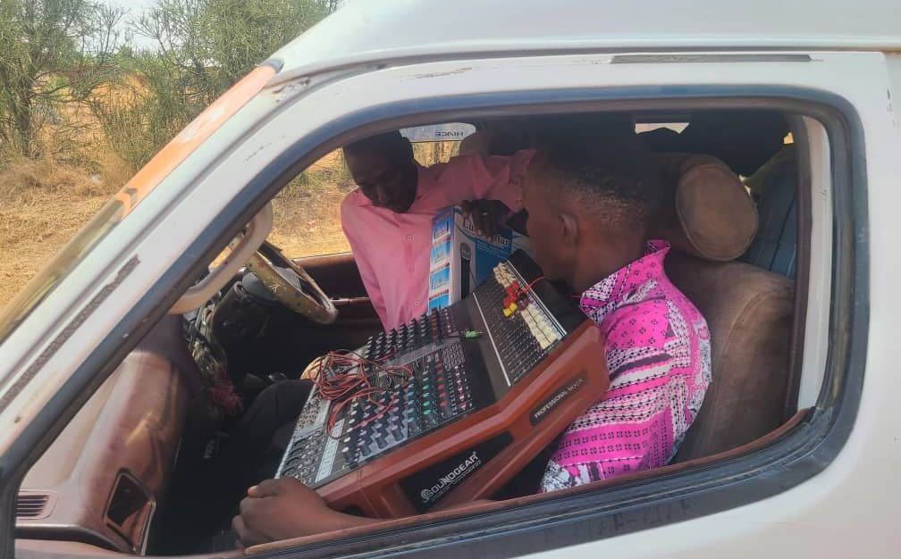 A man in a pink shirt is sitting in a car holding a mixer.