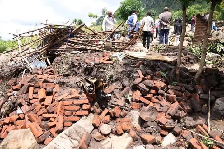 People stand near the ruins of a collapsed brick building in a grassy, mountainous area.