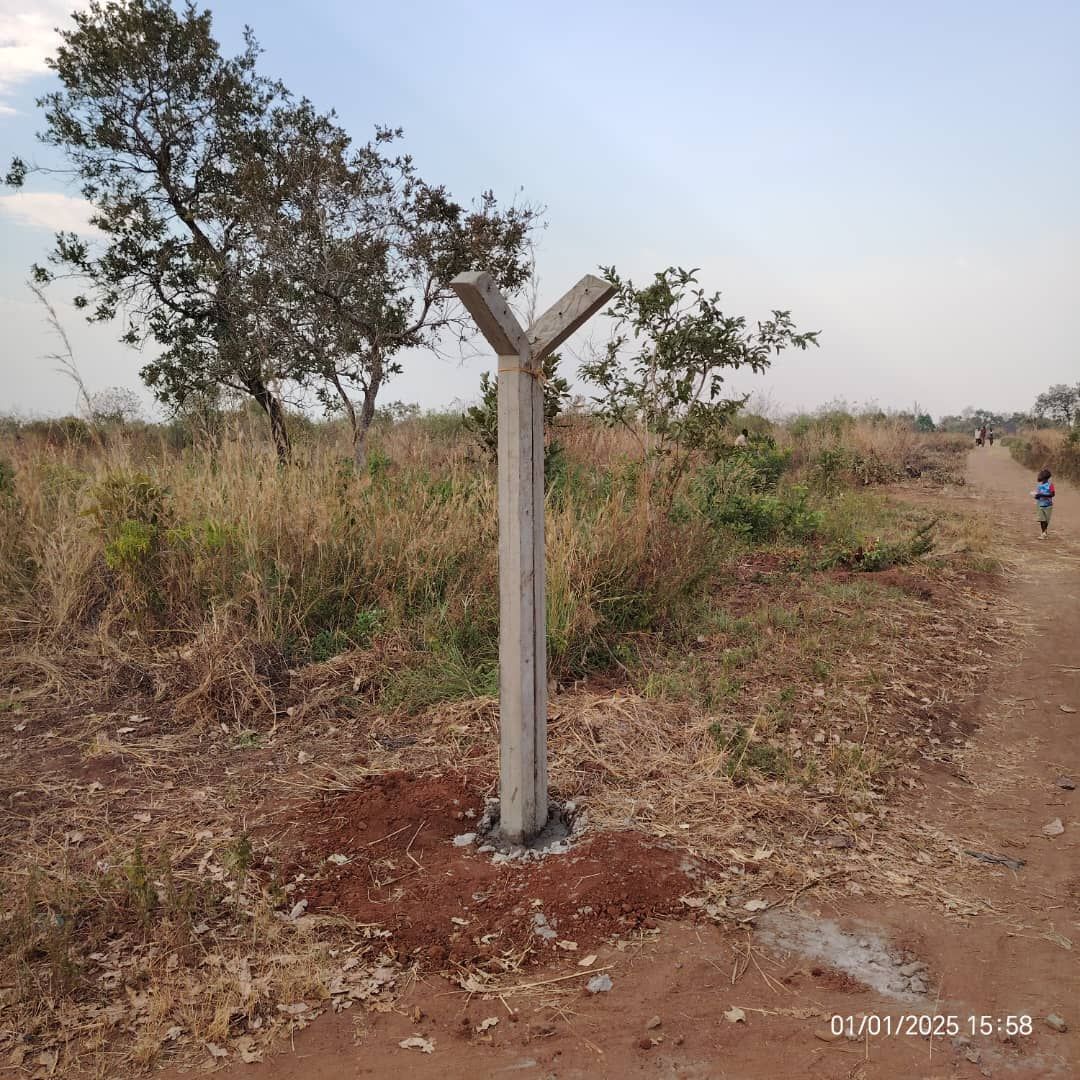 Concrete post with three arms stands in a grassy field next to a dirt path. A person walks in the distance.