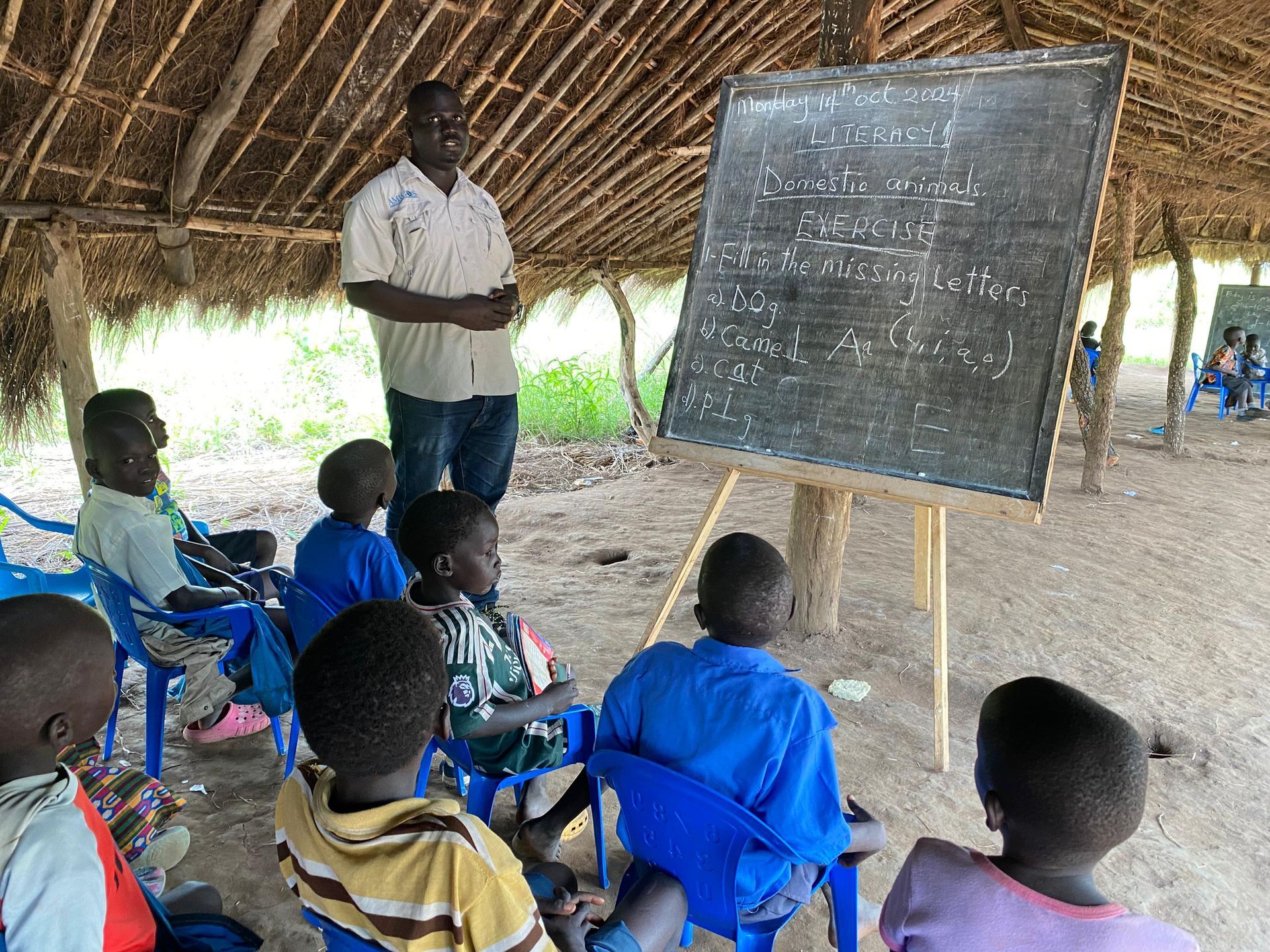 A group of children are sitting under a thatched roof in front of a blackboard.