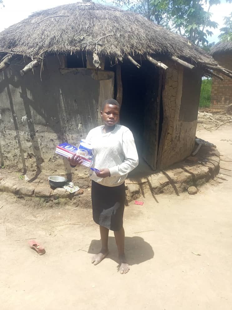 A sponsored child is standing in front of a thatched hut holding a box.