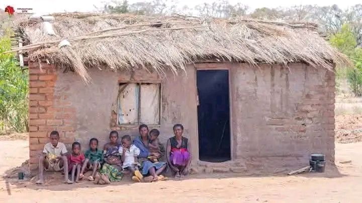 A family of eight sits outside a small, mud-brick house with a thatched roof, in a dry, rural setting.