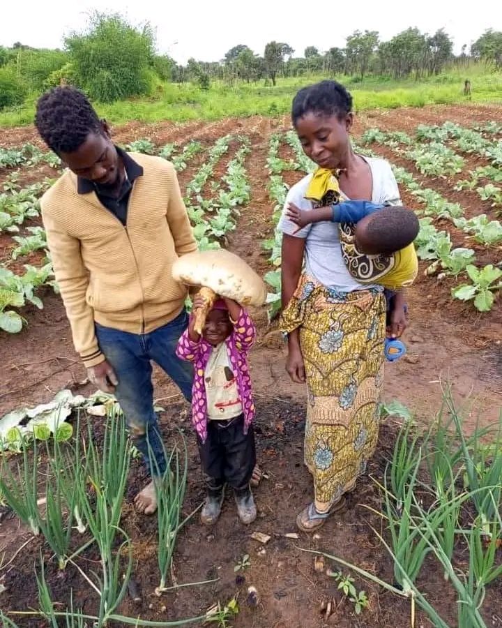 Family in a field; child holds giant mushroom. Parents smile. Lush crops surround them.