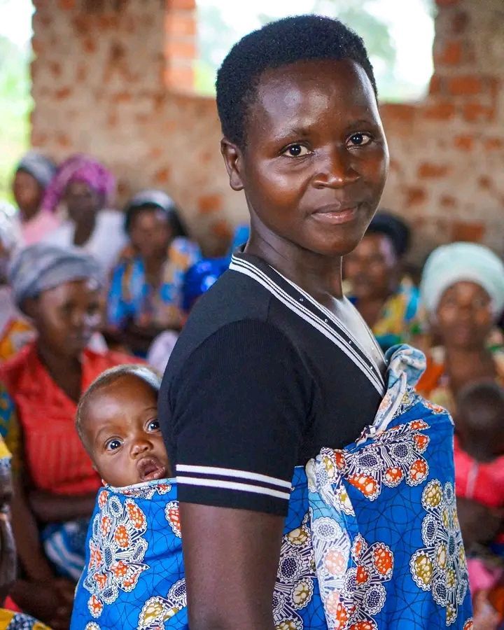 Woman carrying a baby in a blue wrap. Smiling, standing in a room with other women.