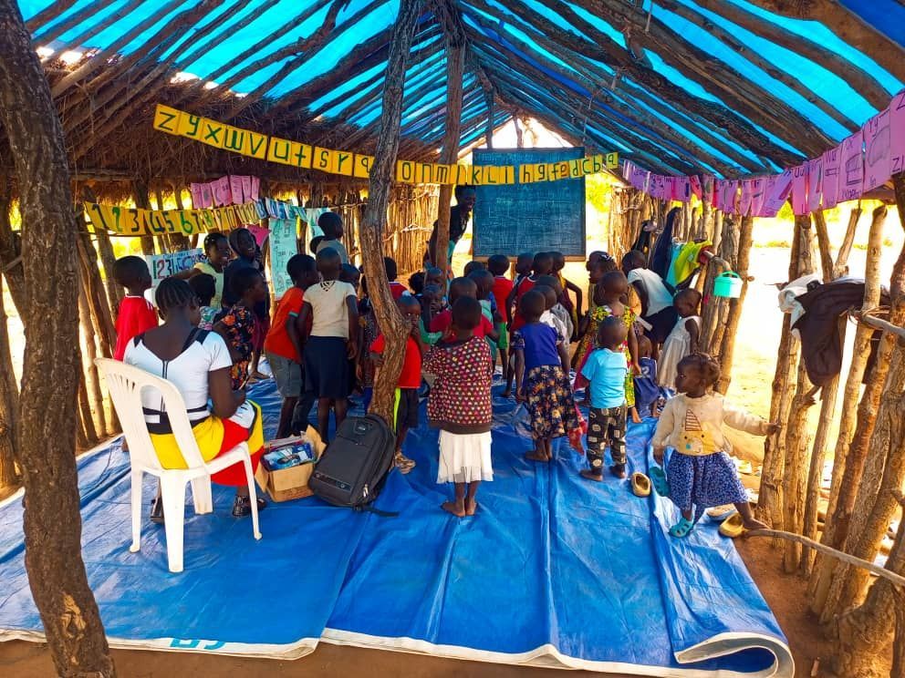Pogo, Northern Uganda — a teacher at the blackboard teaching numbers to dozens of children seated on blue tarps inside the thatched-roof Acema Baptist Church-school, August 2025.