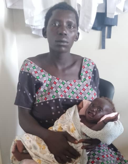 Woman holding infant, both looking at the camera. The woman wears a printed dress, seated indoors.