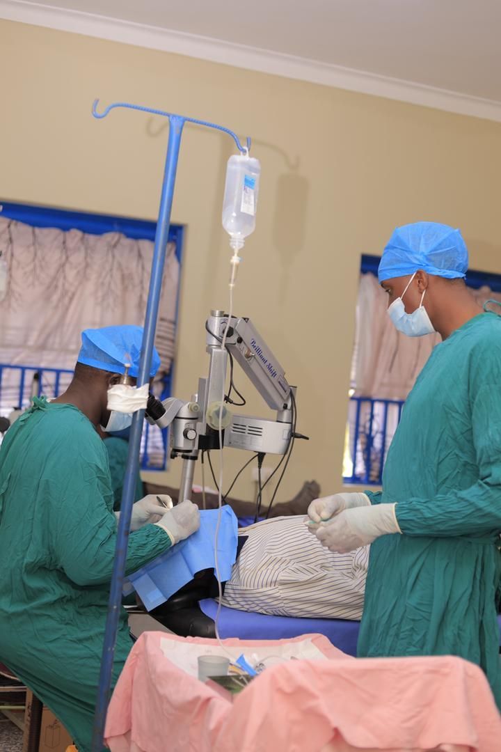 Surgeons in green scrubs operate on a patient under a surgical light, with an IV drip running.