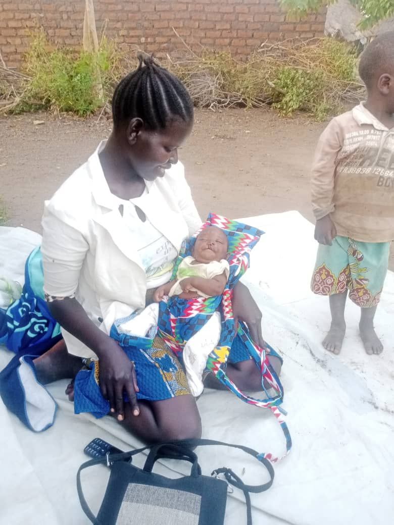Woman seated, cradling a baby swaddled in colorful fabric. A small child stands nearby.