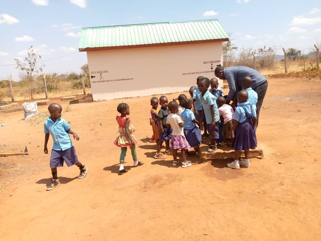 Children in school uniforms gather outside a building with a teacher on a sunny day.