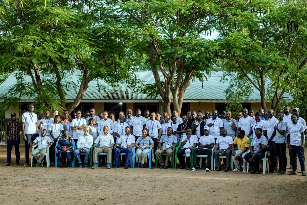 Large group posing outdoors under trees, with many seated and standing in front of a building.