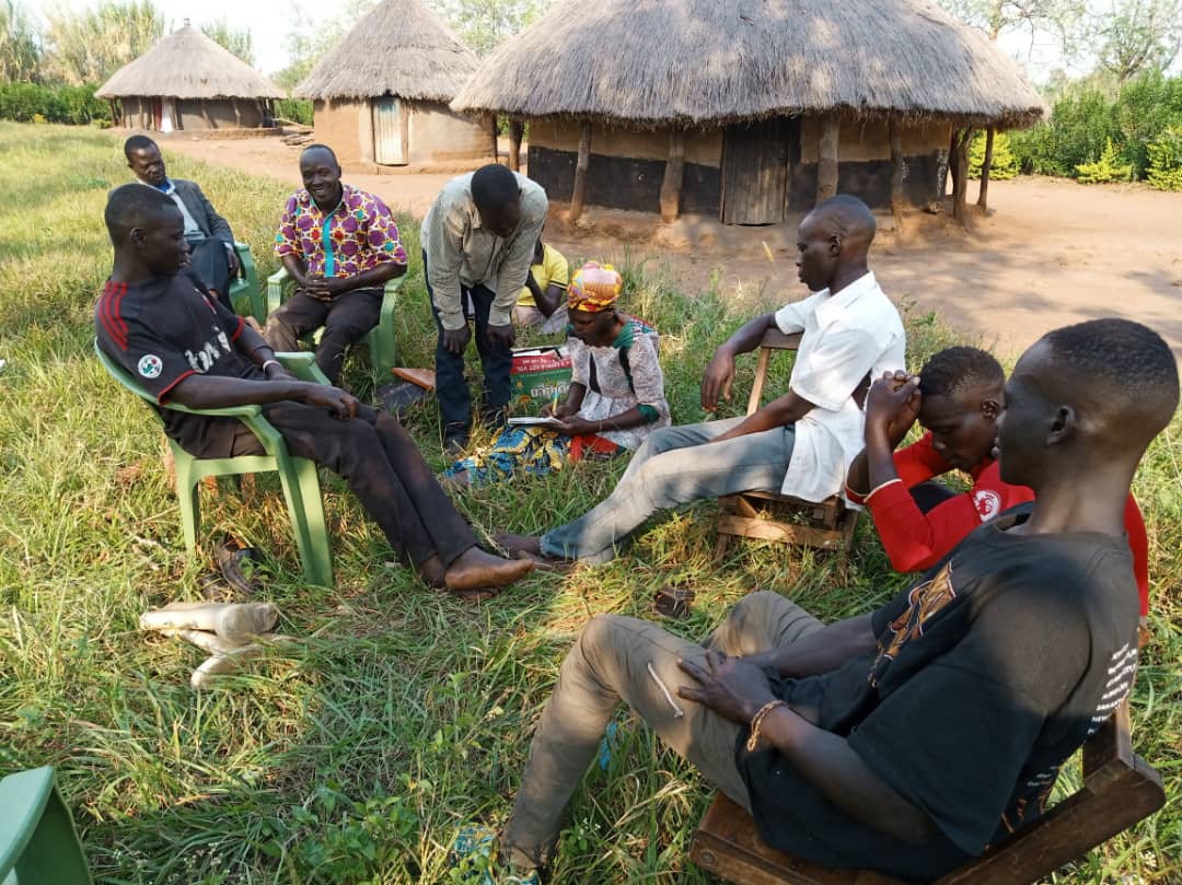 A group of men are sitting in a circle in the grass in front of a thatched hut.