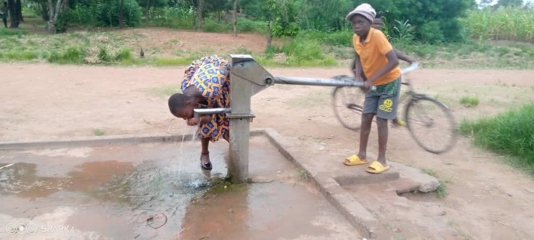 Person stands by a roadside hand pump, filling a bicycle with water beside a muddy puddle.