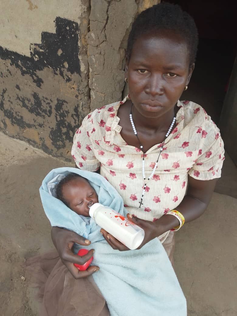 Woman holds and feeds a baby from a bottle. They are outside a building; woman looks concerned.