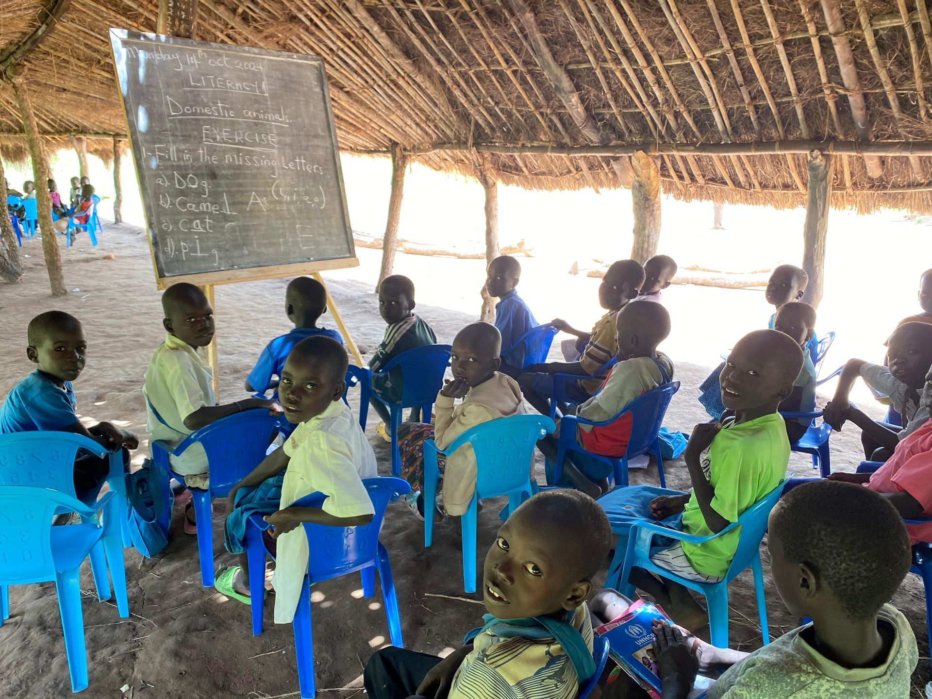 A group of children are sitting in a classroom under a thatched roof.