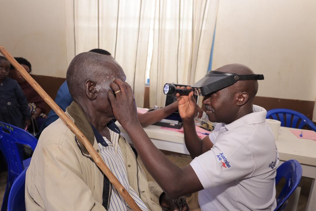 Doctor examining an elderly man's eye. Doctor wears headlamp, and the patient holds a walking stick indoors.