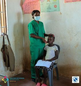 A healthcare worker in green scrubs examines a child sitting in a room.