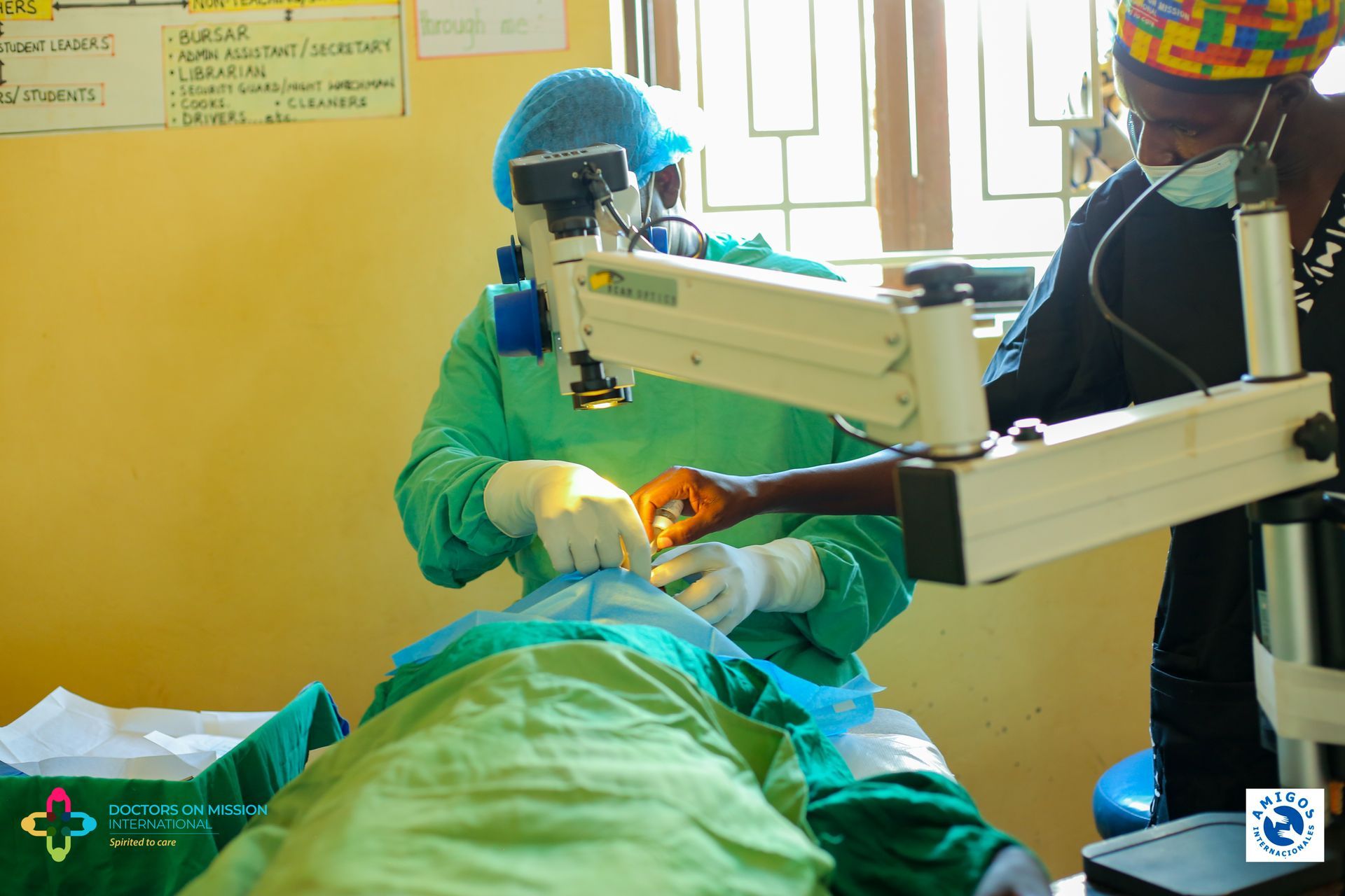Surgeon in green scrubs and goggles performs eye surgery with assistance. Yellow walls, bright sunlight.