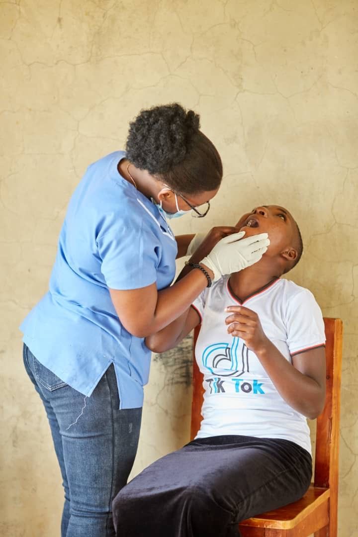 A dental professional examines a patient's mouth. Both are Black. Patient is sitting, expressing concern.