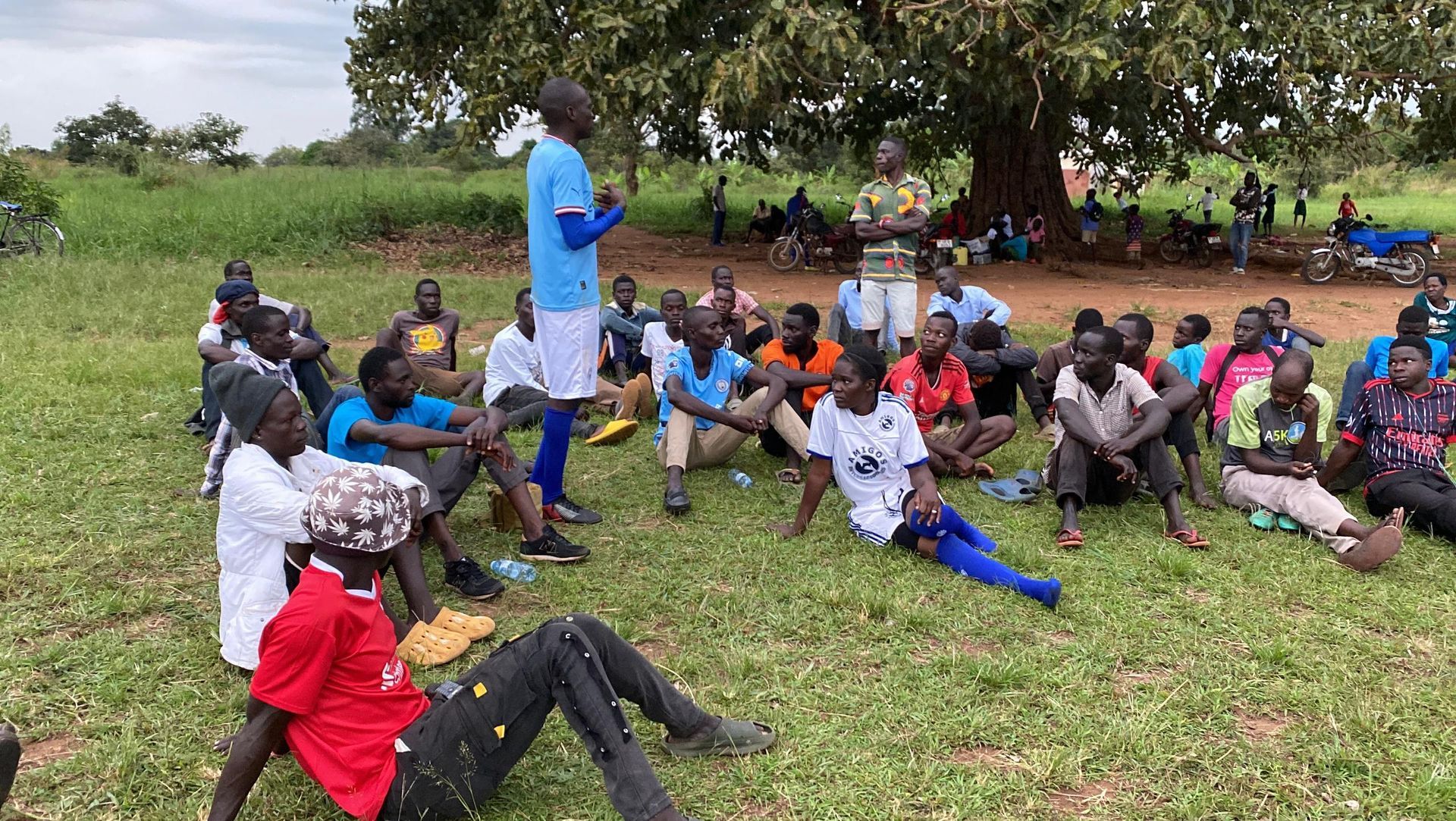 A man is talking to a group of people sitting on the grass.