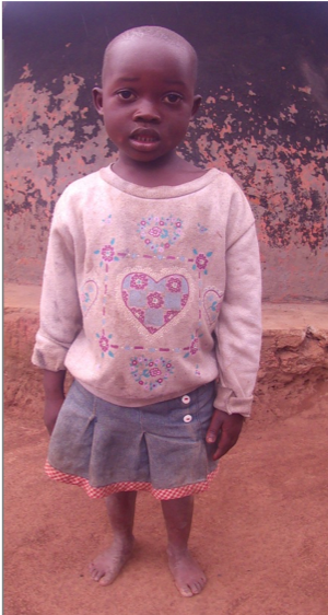 Young child with shaved head, standing, wearing light-colored sweater and denim skirt; outdoor setting.