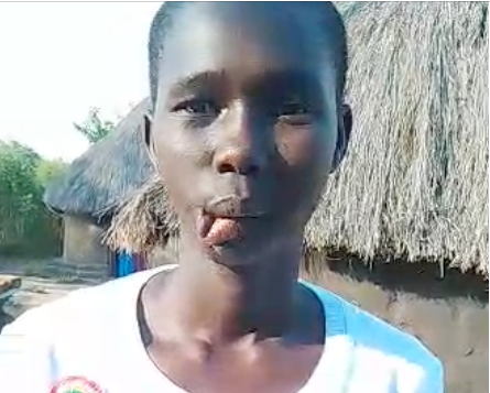Young lady outdoors, slightly sticking out tongue. Background has thatched roof building and trees.