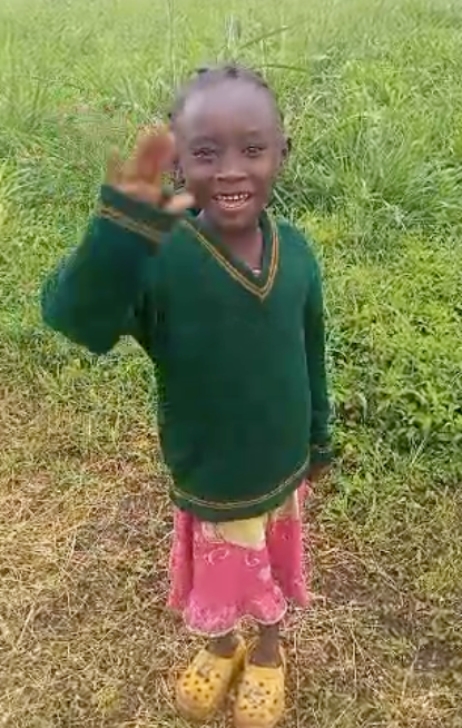 Child waving, smiling, wearing green sweater and pink skirt, standing in a grassy field.
