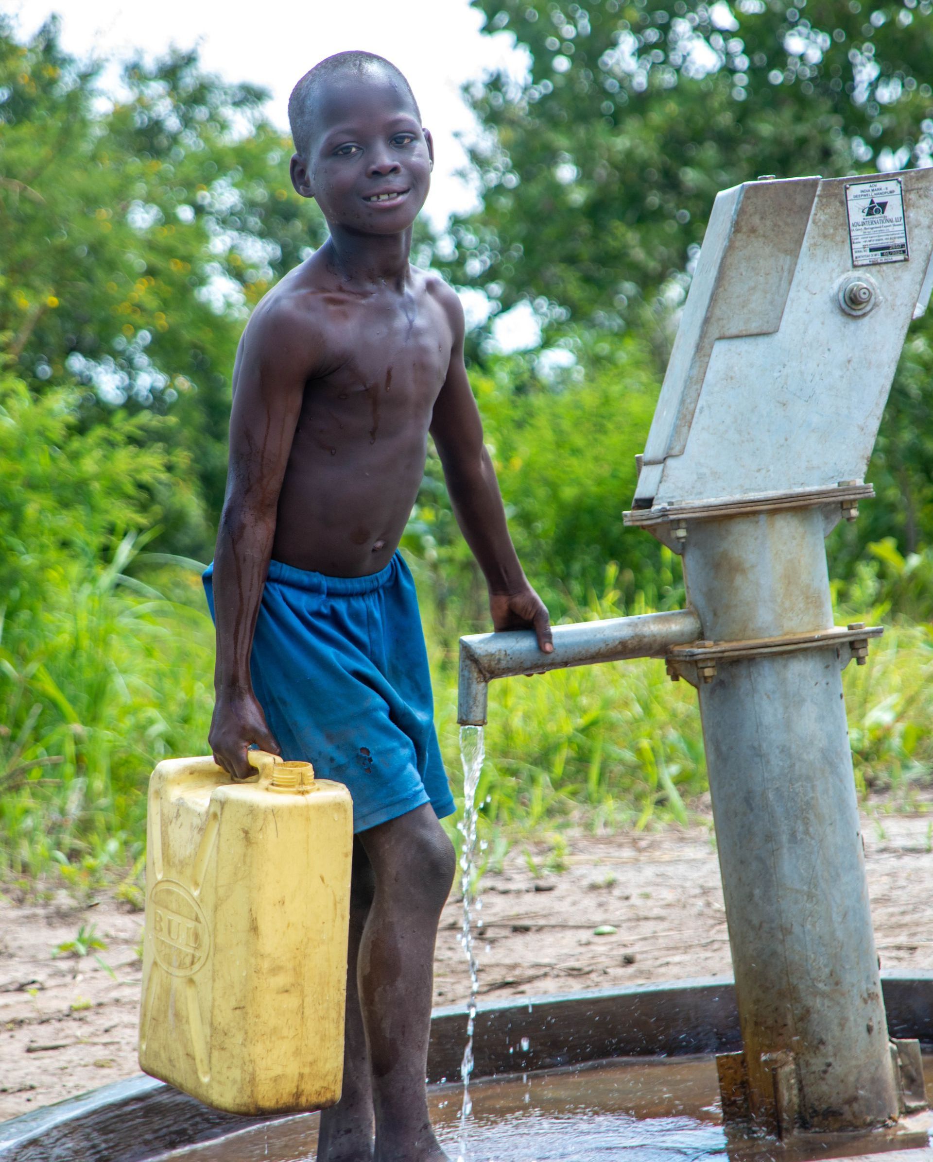 A young boy is standing next to a water pump holding a yellow container