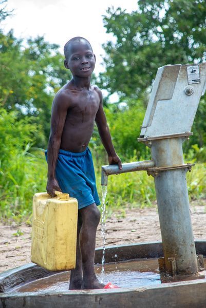 Boy pumps water from a well, filling a yellow jug. Smiling, outdoors in a rural area.
