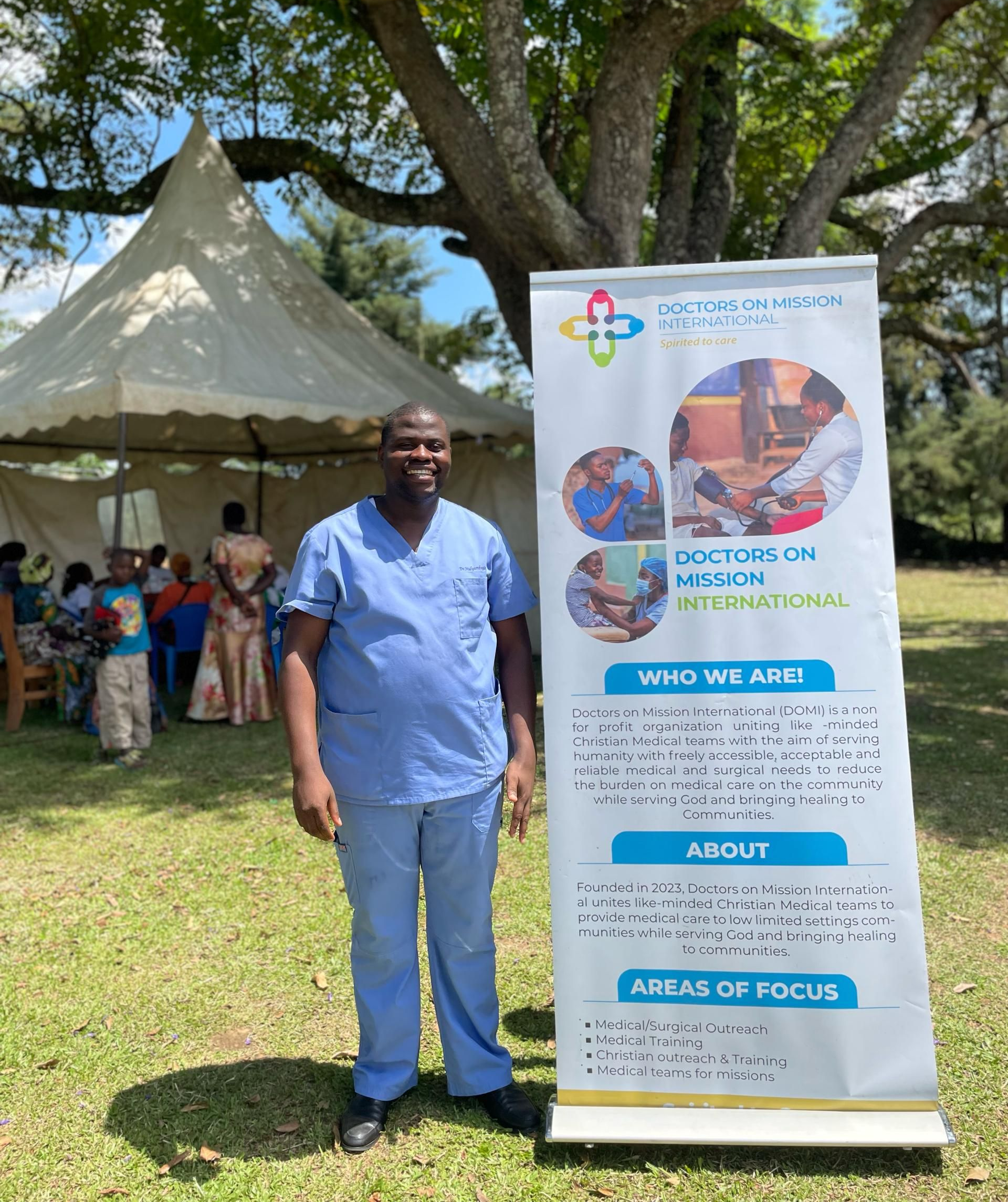 Man in blue scrubs stands by banner in a grassy outdoor setting.