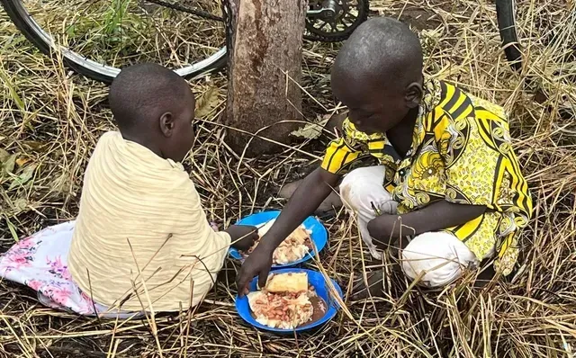 Two children eating food from blue plates outdoors near a tree, in dry grass.